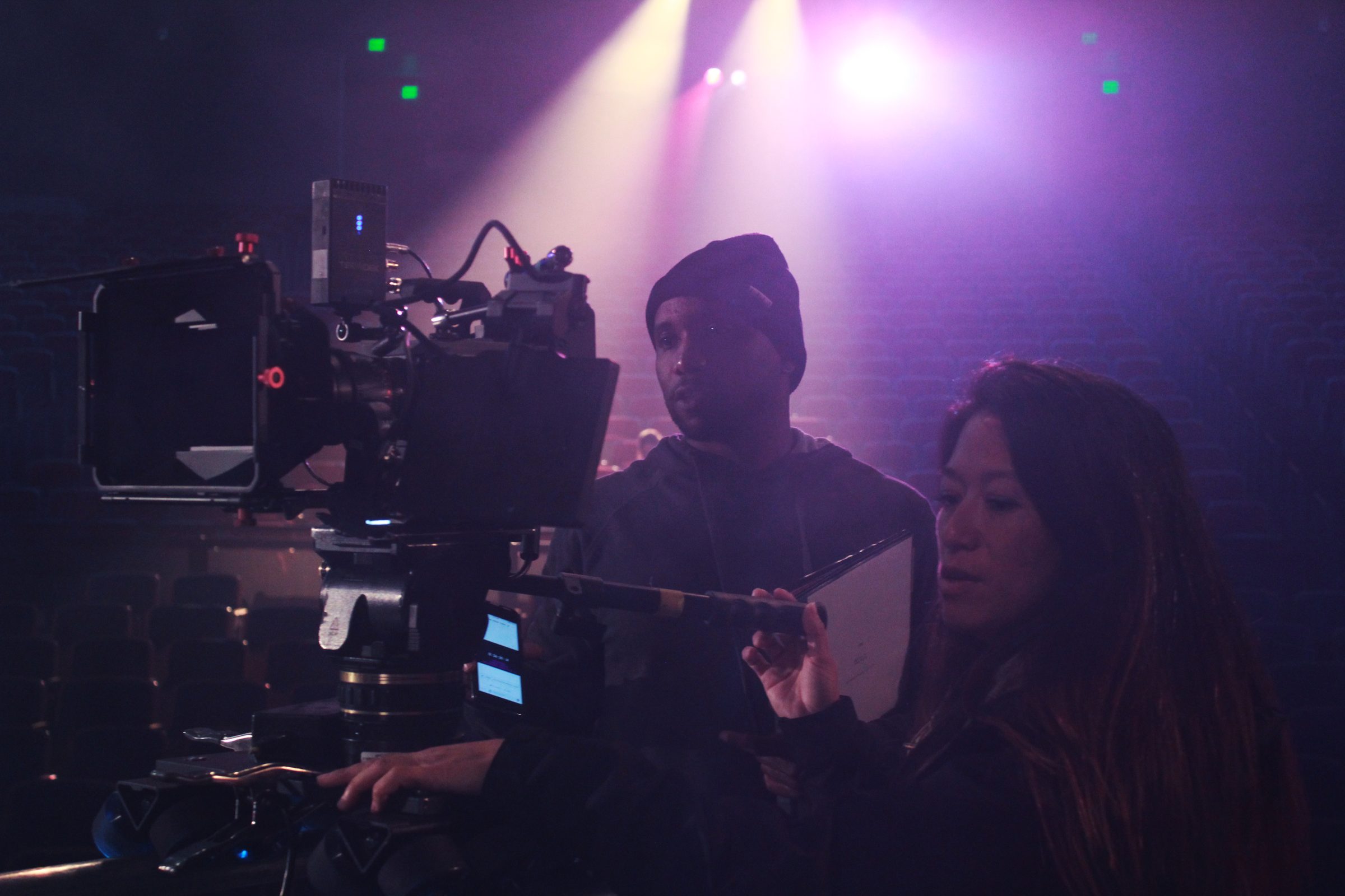 Isaiah beside the camera with the operator in a theater lit by stage lights